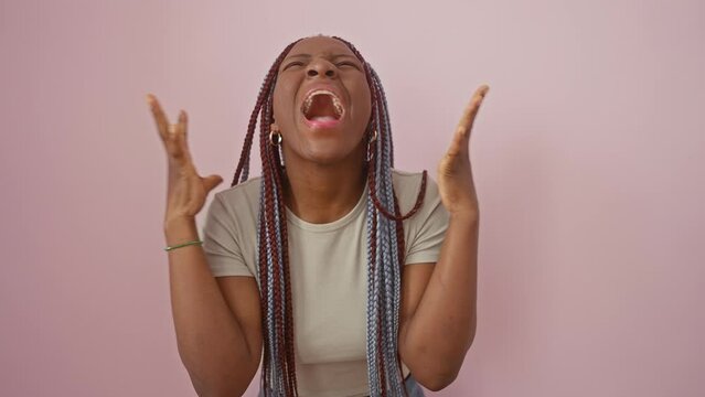 Stressed-out african american woman lets loose with angry shouts, yelling in a frenzy, arms raised high in rage. mad expression of frustration on pink isolated background
