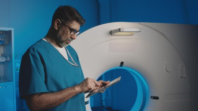 Medium slowmo shot of male multiethnic radiologist in uniform holding tablet, looking at MRI machine and double-checking MRI examination appointments