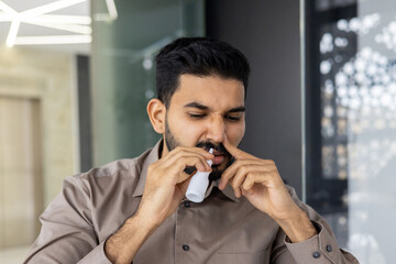 A close-up photo of a young Indian man who is being treated while in the office at the workplace, blowing his nose with medication for a sore throat.