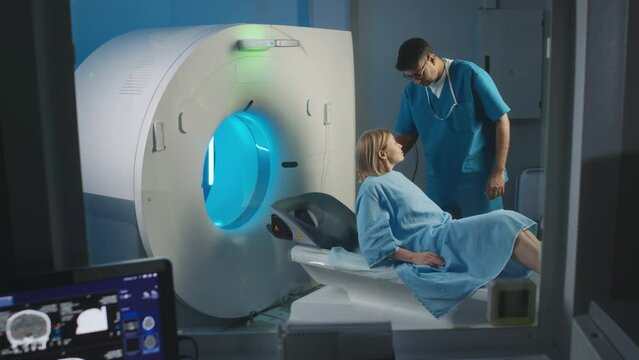 Medium slowmo shot of mature Caucasian woman in hospital gown lying into MRI capsule under supervision of male radiology technician holding remote control and explaining diagnostic imaging procedure