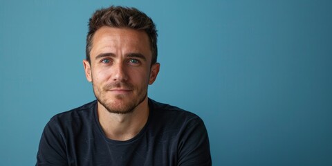 Fototapeta premium Young man with short hair and a black shirt against a blue background, looking confident.