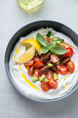 Bowl of greek feta dip topped with fresh tomatoes and cucumber, vertical shot on a light-grey granite surface, elevated view