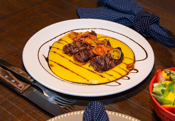 White dish with soft polenta, sliced ​​meat on top of the polenta and tomato confit with sauce on top of the meat. Wooden table ready to eat with blue fabric napkin and cutlery on it.