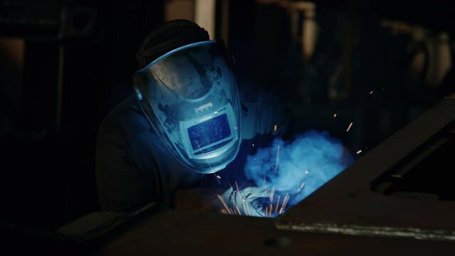 View from above of metalworker in safety mask welding iron details with torch producing sparks in industrial factory
