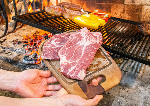 Chef hand holding raw meat on a wooden board in front of a Uruguayan parrilla fire. Roast meat being made in a restaurant kitchen.