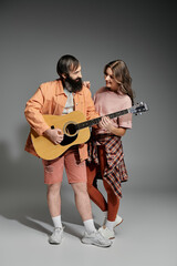 A couple poses together in a studio, the man playing a guitar while the woman leans in and smiles.