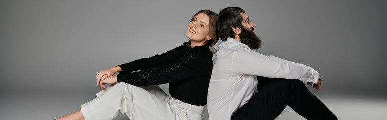 A loving couple sits back-to-back, dressed in sophisticated attire against a grey background.