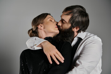 A couple dressed in sophisticated attire share a passionate kiss against a simple gray background.