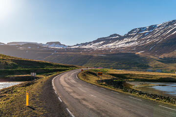 Road towards Ísafjörður, with the town sign. Mountains with snow, bright sunshine, early summer. Green grass and sea