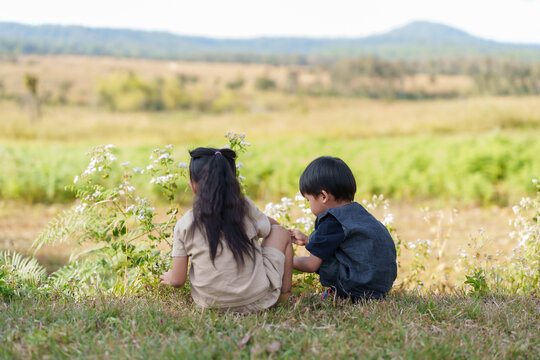 Young boy and girl sitting on grass in national park, exploring flowers. Captures innocence, curiosity, peaceful nature of family vacation. Sunny day, green fields, mountains in distance.