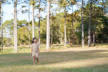 Naklejka premium Young girl in beige outfit walking in national park, wearing blue headphones, smiling. Captures joy and energy of family vacation in nature. Bright sunny day, green trees in background.