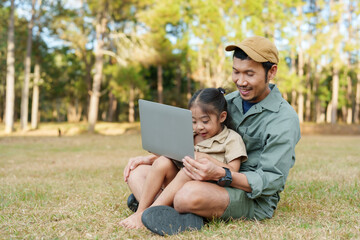 Father and young daughter in national park, using laptop together, smiling and bonding. Captures joy of family vacation in nature. Bright sunny day, green trees in background.