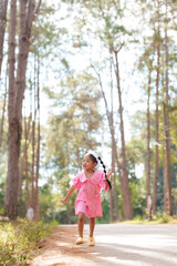 Asian girl in pink dress running joyfully along forest path. Smiling, arms outstretched, embracing nature. Sunlight filters through trees, creating vibrant, happy atmosphere.