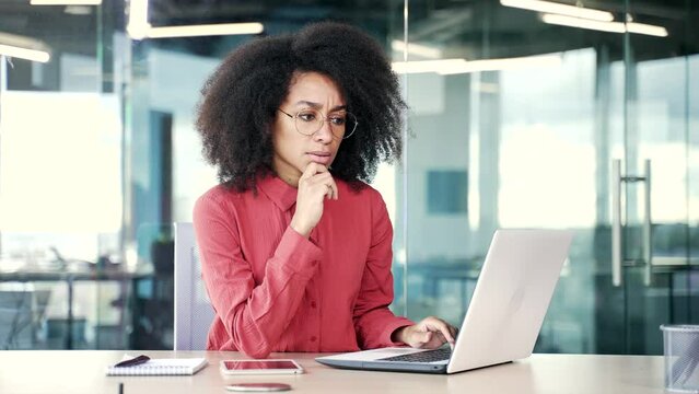 Thoughtful concentrated young african american female employee typing on laptop and thinking about problem solving at workplace in office. Serious black woman is working in computer application - Powered by Adobe