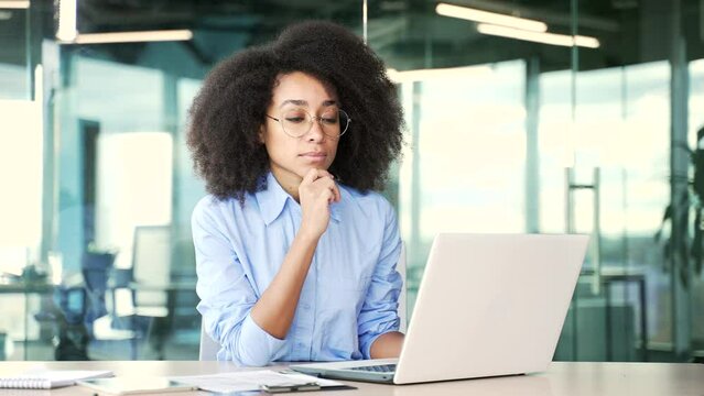 Thoughtful focused young african american female employee typing on laptop and thinking about problem solving at workplace in office. Serious black woman is working in computer or texting a client