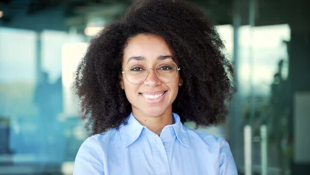 Portrait of a young happy african american female employee in a blue shirt standing in business office. Smiling black woman in glasses posing looking at camera. Headshot of a positive worker. Close up