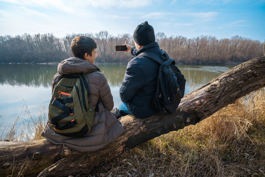 Two men sitting on a log by the river, taking photos on a smartphone, father and son, early spring landscape, the concept of hiking and outdoor activities