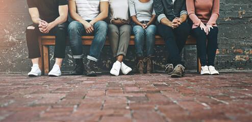 Creative people, legs and waiting room with career opportunity, interview or startup on brick wall. Low angle of group, team or community sitting on outdoor bench for job, line or row together