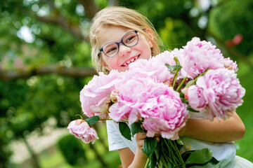 Fototapeta premium portrait of a happy cute little caucasian seven year old kid girl, holds in hands and smell and enjoying a bouquet of pink peony flowers in full bloom on the background of nature