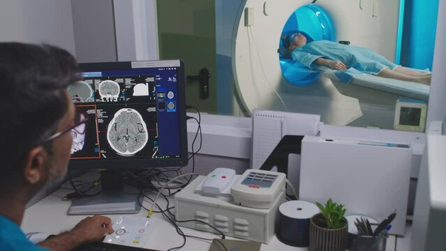 Back view of multiethnic male medical imaging specialist sitting in MRI room, controlling and operating scanner. Female patient in hospital gown undergoing diagnostic procedure, lying on patient bed