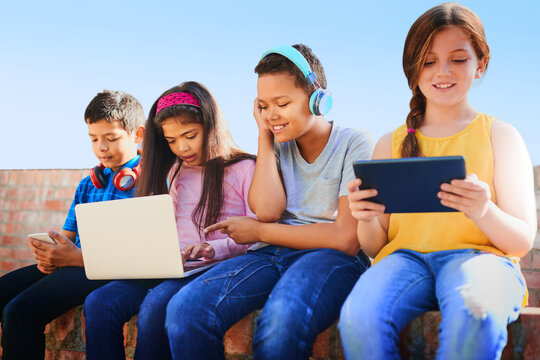 School kids, friends and happy with phone, tablet and laptop with diversity, relax and streaming in summer. Girl, boy and group with headphones, smile and games with subscription on wall at academy