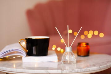 Cup of tea with open paper book and liquid home diffuser in glass bottle and bamboo sticks staying on marble coffee table close up. Cozy hygge atmosphere.
