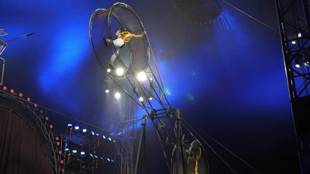 Brave acrobats perform tricks on the spinning wheel of death under the dome of the circus. A huge spinning wheel with acrobats inside, acrobats jumping and tumbling. A circus show.
