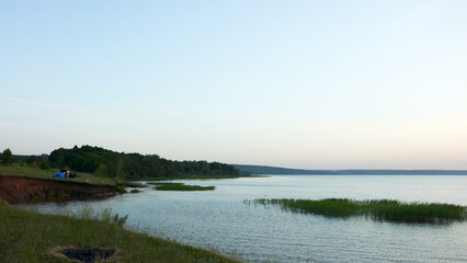 Wild camping on a picturesque riverbank during sunset. A car and a tourist tent near the shoreline.  There are reeds in the water.