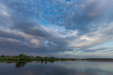 Calm waters reflect the pastel hues of the sky as fluffy clouds drift overhead, capturing the peaceful essence of a tranquil spring evening by the lake.
