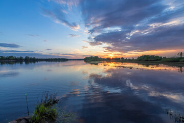 A captivating image of a calm river reflecting the vibrant hues of a spring sunset, with a distant shoreline and a peaceful fishing rod in the foreground.