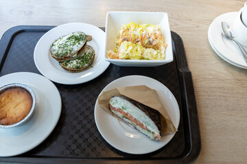 A close-up of a lunch tray with a variety of food items. There are fluffy pancakes topped with herbs, a creamy salad with vegetables and sauce, and a delicious-looking sandwich wrapped in paper.