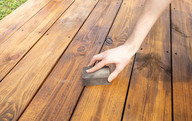 A close-up shot of a hand sanding a wooden deck with a block of sandpaper. The deck is made of...