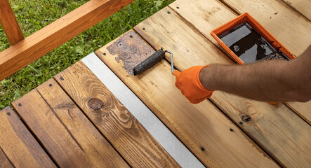 A close-up shot of a hand wearing an orange glove applying a dark stain to a wooden deck using a paint roller.