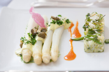Elegant white asparagus dish with garnishes, accompanied by a side and decorative sauces on a plate.