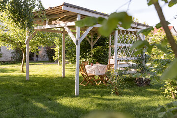 White gazebo with wooden furniture in garden. Summer background. Beautiful landscape of a country house.