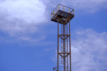 the observation tower at the factory against the background of the blue sky with CCTV cameras. A narrow iron ladder for lifting an employee to the tower.