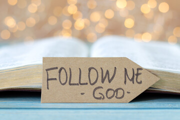 Follow Me, God, handwritten text on paper arrow with open holy bible book and bokeh light...