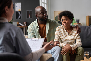 Portrait of teenage boy listening to psychologist during family therapy session with caring father...