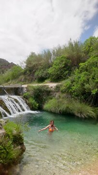 A woman is bathing in a stream. The water is clear and the surrounding area is lush and green