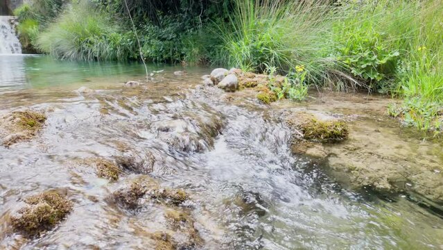 A stream of water flows over rocks and through a grassy area. The water is clear and calm, and the surrounding vegetation is lush and green. The scene is peaceful and serene