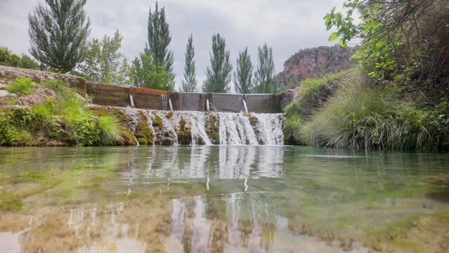 A beautiful stream of water flows through a lush green forest. The water is crystal clear and the trees are tall and green. The scene is peaceful and serene