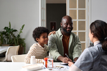 Portrait of smiling African American father and son signing medical form with female doctor giving...