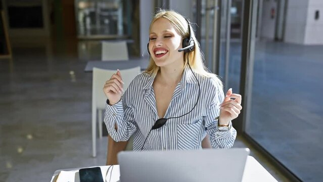 A cheerful blonde woman wearing headphones works on her laptop at a modern office table.