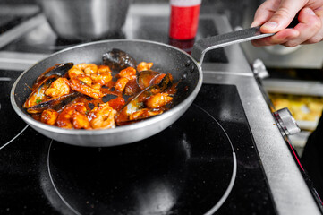 Close-up view of a pan on a stove, with sautéed shrimp and vegetables being stirred by a person...