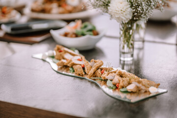 Valmiera, Latvia - September 9, 2023 - Close-up of a plated dish with breaded chicken strips and garnish, served on a reflective platter with a floral centerpiece in the background.