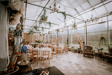 Valmiera, Latvia - September 9, 2023 - Elegant wedding reception setup in a greenhouse with round tables, floral arrangements, and hanging greenery, creating a charming atmosphere.