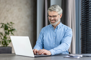 Fototapeta premium Elderly man with glasses and a beard smiling while using a laptop in a modern office environment