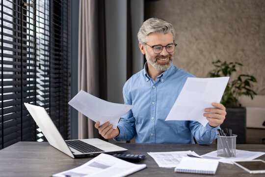 Confident businessman smiling while reviewing documents at his desk with a laptop in a modern office. Professional setting with natural light and organized workspace.