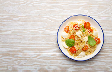 Top view of a plate of spaghetti pasta with cherry tomatoes and mozzarella cheese on a white wooden background with copy space