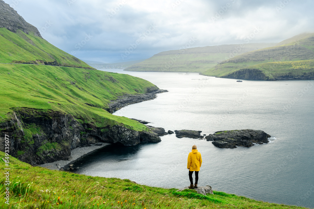 Wall mural lonely tourist standing on edge of the cliff on vagar island and sorvagur town on background. faroe 